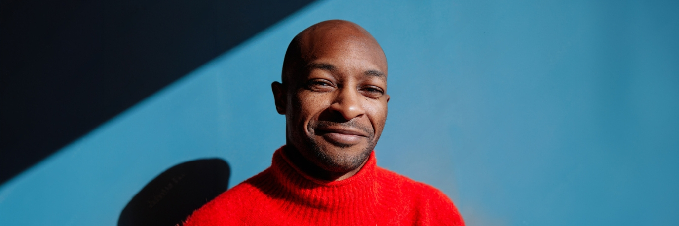 Man of color wearing a red pullover and smiling in front of a blue background